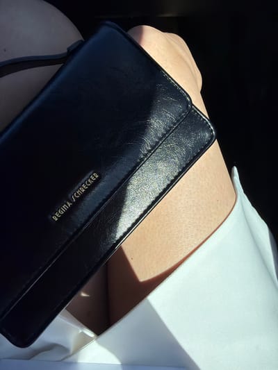 A black leather purse with "Regina Schrecker" embossed in gold lettering sits in the lap of a person wearing a white collared shirt. The purse is angled towards the viewer, with the text oriented horizontally. The person's skin is light-toned. The location is not identifiable as Manole, Bulgaria, based on the visual information provided.