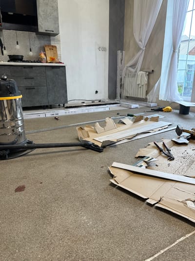 This is a photograph of an interior scene that appears to be under renovation or construction. The subjects include building materials such as long, flat pieces of wood or laminate flooring, cardboard boxes, and what looks like part of a disassembled kitchen cabinet. There is also a wet/dry vacuum cleaner in the foreground. The setting is a room with a concrete floor. 

In the background, there is a window with sheer white curtains, a radiator, and what could be kitchen cabinets or built-in storage units. The walls are painted in neutral tones, with one section showing a geometric patterned wallpaper or tile. There are some tools visible on the floor, including pliers and a hammer. A yellow object, possibly a piece of safety equipment like a hard hat, is also present. 

The overall impression is one of a work in progress, with materials and tools scattered as if someone was actively working on a project. There are no people visible in the image. The lighting suggests it is daytime, but the weather cannot be determined. 

There are no prominent text elements visible in the image. The location is identified as Kuyuki, Russia.