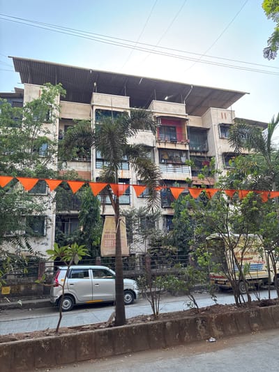 The image captures a street scene in Ambernath, India. In the foreground, a parked silver hatchback car is visible. The road and a raised curb are also present. Behind the car, a multi-story residential building stands, partially obscured by trees and a string of orange triangular flags. The building's facade shows signs of age and has a protective metal awning on the top. Windows are visible, some with security bars or enclosed balconies. A delivery truck is parked along the road further down. Power lines are visible in the upper left corner against a bright sky.
