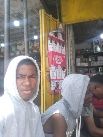 The image depicts three Black males in a commercial setting in Igwuruta, Nigeria. In the foreground, an individual wearing a light grey hooded sweatshirt with the hood up stares directly at the camera with a furrowed brow. To his immediate right and slightly behind, a second male wears a grey hooded sleeveless top with the hood up and looks towards the right. A third male, partially visible on the far right, wears a dark shirt. The background features a storefront entrance characterized by a yellow-painted metal structure. A red and white advertising banner, displaying product images and the text "STAY INSPIRED," is affixed to this yellow frame. Within the visible shop interior, shelves are stocked with various electronic items including headphones, speakers, and boxed goods. Overhead, multiple white LED light fixtures illuminate a light-colored, patterned ceiling.