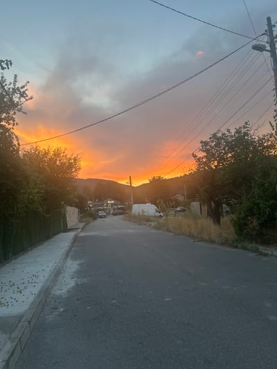 This media file captures a dramatic sunset over Boboshevo, Bulgaria. The scene is dominated by the sky, painted in vibrant hues of orange and yellow, suggesting the golden hour or a vibrant twilight. A mountain range forms a dark silhouette against the bright sky. In the foreground, a paved road runs along the right side of the frame, with a concrete sidewalk and curb. On the left, a utility pole with several power lines stretches across the sky. Trees and vegetation line the roadside and the base of the mountain. Several buildings and parked vehicles are visible in the mid-ground, indicating a residential or commercial area. The overall impression is one of natural beauty and a peaceful evening setting. There is no discernible text in the image.