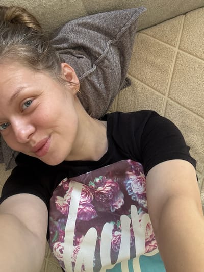 An overhead, close-up, selfie-style shot shows a woman lying down on a textured, light brown, tiled surface, with a dark grey, textured cushion behind her head. She is wearing a black t-shirt with a floral pattern of pink and purple roses and white lettering on the chest. Her hair is pulled up in a bun on the top of her head, and she is looking towards the camera with a slight smile. Her left ear is visible, adorned with a small gold hoop earring. A silver nose ring is also visible on her left nostril. The lighting is natural and appears to be from above, creating soft shadows.