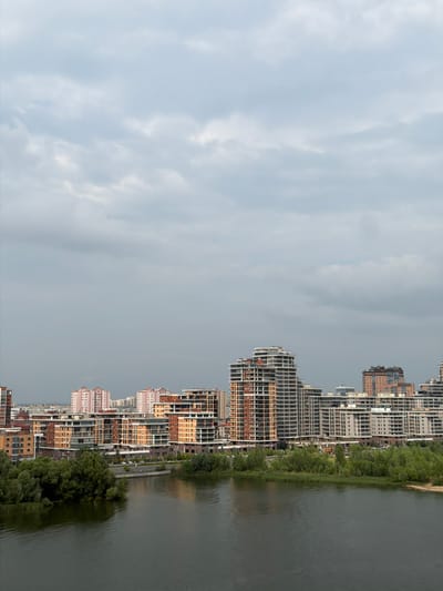 This is a wide, horizontal, aerial shot of a modern city skyline situated along the bank of a dark, tranquil body of water. The sky is overcast with a thick blanket of gray clouds, contributing to a muted, subdued atmosphere. In the foreground, the water reflects the overcast sky, creating a dark, rippling surface.
Along the far bank, a dense line of lush green trees forms a verdant barrier between the water and the buildings. Behind the trees, a collection of modern, multi-story buildings rises, displaying a variety of architectural styles and colors. Some buildings are clad in warm, earthy tones of orange and brown, while others feature lighter, neutral palettes of white and gray. Several buildings exhibit complex, geometric designs with numerous balconies and windowed facades.
Further into the distance, additional buildings of varying heights and designs are visible, creating a layered urban landscape. The overall impression is of a developing city with a significant emphasis on modern residential and commercial construction. There are no visible people or specific activities captured in this scene. The location is identified as Tarlashi, Russia.