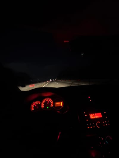 The image shows the interior of a car at night. The dashboard and speedometer are visible, illuminated in red. The speedometer indicates a speed of approximately 100 km/h. The radio display is also illuminated, showing text in an unfamiliar script. Through the windshield, a dark road with faint lights in the distance can be seen. It is night. The location is in the city of Trilistnik, Bulgaria.