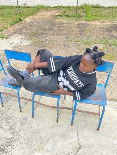 A young Black individual with their hair styled in multiple bantu knots is lying down on two attached blue metal chairs. They are wearing a black long-sleeved shirt with white stripes on the sleeves and white lettering on the front that reads "FERRIPAYS." They are also wearing loose-fitting, dark-wash denim shorts and black platform loafers with socks. A beaded bracelet with a cross charm is visible on their left wrist. The chairs are situated on a cracked concrete surface, with a chain visible draped over the individual's lap. In the background, there is a chain-link fence, green grass, and some debris. The scene appears to be outdoors, possibly in a park or schoolyard, in the city of Zawan, Nigeria.