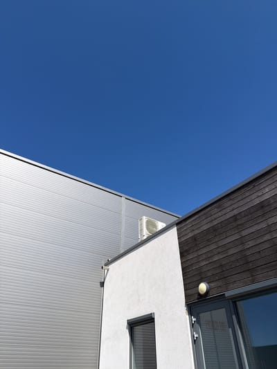 The image shows an abstract view of a modern building with a clear blue sky. The building features a dark wooden facade at the top, which angles downwards towards a textured white section with a window. Below this, a large, ribbed silver metal facade extends downwards and to the left. A small, circular outdoor light is visible on the underside of the wooden section. An air conditioning unit is mounted on the white part of the building, partially obscured by the angled wooden structure. A simple gutter pipe extends from the white section. The sunlight suggests it is daytime, and the clear sky indicates fair weather. The angle of the shot, looking upwards, emphasizes the geometric lines and contrasting materials of the architecture against the vast expanse of the sky. No people or specific events are depicted.