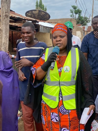 In an outdoor scene in Rigachikun, Nigeria, a woman wearing a black hijab, an orange and purple patterned dress, and a bright yellow safety vest is speaking into a blue microphone. She is holding a stack of white papers in her right hand. The vest has a logo with the words "NATIONAL ORIENTATION AGENCY NOA" printed on it. To her left, a young man in a blue and white striped t-shirt looks towards her, smiling. Behind the woman, another man wearing a green cap and a white shirt is visible. In the background, there are structures with corrugated metal roofs, some appearing worn, and electrical wires running overhead. The ground is uneven and appears to be a dirt path with scattered debris. A purple hooded garment is visible on the far left, suggesting other individuals are present. The overall setting appears to be a modest urban or semi-urban environment.