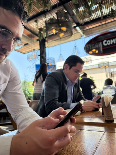 The image depicts an outdoor or semi-outdoor restaurant or cafe setting within a modern commercial complex or shopping mall in Santiago de Querétaro, Mexico.

In the foreground, a light-skinned male wearing round framed glasses and a white long-sleeved shirt is seen from the chest up, looking down at a dark smartphone held in both hands. His right hand is prominently visible, operating the device. To his right, in the mid-ground, another light-skinned male wearing glasses, a light blue collared shirt, and a gray plaid suit jacket is similarly engrossed in a dark smartphone, held with both hands on a wooden table. A small wooden napkin holder with white napkins sits on the table to his right.

The environment features a prominent slatted wooden ceiling structure adorned with hanging greenery and numerous decorative light fixtures. These fixtures are predominantly dark metal cages containing multiple warm-toned Edison-style bulbs, with some smaller, simpler geometric cages also visible. The background shows a clear blue sky through large openings or windows, indicating bright daylight. Out of focus, several other individuals are present: a person in a white striped top walking away in the background to the left, and a dark-clad individual further back to the right. Signs are partially visible in the distance, including a red-rimmed black sign with white text partially reading "STEAK COMBO" and another sign displaying Asian characters. The overall scene suggests a casual yet somewhat upscale dining experience.