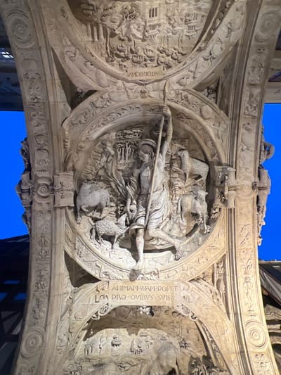 The image displays a brightly illuminated, ornate stone architectural relief, likely part of a portal or archway, set against a deep blue background, indicating a night scene with artificial lighting. The location is in the city of Rouen, France.

The central feature is a large circular medallion carved in high relief, depicting a male figure identified as the Good Shepherd. He is shown with one leg bent and the other extended, holding a long staff or crook diagonally across his body with his left arm raised. He wears a simple tunic and sandals. Several sheep are carved around him within the medallion, some standing, some looking towards him. The background within the medallion shows carved trees and foliage, indicating a pastoral setting.

Below this central medallion, a rectangular panel bears a Latin inscription in capitals: "ANIMAM SVAM DONAT PRO OVIBVS SVIS" (He lays down his life for his sheep).

Above the central medallion, another arched section features a different narrative relief with numerous smaller human figures engaged in various activities, and partially visible text that appears to read "PASTOR BO" (likely part of "Pastor Bonus").

The entire structure is composed of multiple layers of arches or ribs, all elaborately carved with intricate decorative motifs. These include vertical bands of scrolling patterns, floral elements, geometric designs (such as a concentric circle), and smaller human or figural elements. The stonework is light-colored, showing strong shadows that emphasize the depth and detail of the carvings. Darker architectural elements, possibly rooflines or structural beams, are faintly visible against the blue background at the edges of the frame.