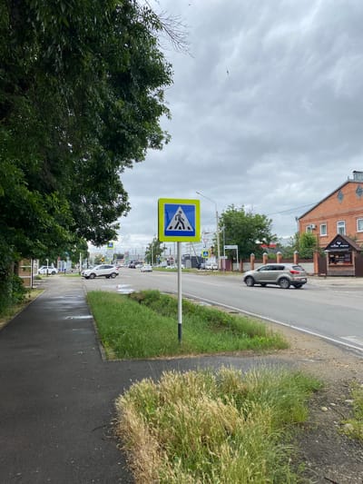 The image shows a street scene in the city of Zapadny, Russia, on a cloudy day. A pedestrian crossing sign is prominently displayed on the right side of the road, featuring a blue square with a yellow border and a black silhouette of a person walking. The sign is mounted on a metal pole. To the left of the sign, a sidewalk is visible, wet from recent rain, with some puddles. Lush green grass lines the edge of the sidewalk and separates it from the road.
The road itself is paved and has at least two lanes of traffic. Several cars are present. A silver SUV is driving on the right side of the road, moving away from the camera. In the background, on the left side of the road, more cars are visible, including a white SUV that appears to be stopped or moving slowly. Further in the distance, traffic lights and other street signs can be seen, along with buildings. One building on the right side of the road is made of red brick and has a small, open-air kiosk with the text "coffee memo" on it. Trees with dark green foliage line the left side of the sidewalk, creating a canopy effect over parts of the scene. The sky is overcast with gray clouds. There are no people clearly visible in the immediate foreground, but there might be pedestrians further down the road near the white SUV.