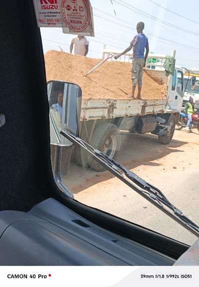 A view from inside a vehicle reveals a truck overloaded with sand. A man stands on the sand bed, holding a shovel. Another person stands beside him on the sand. In the side mirror, another person is visible. The foreground shows the vehicle's interior with the wiper blades visible on the windshield. A sticker with ISUZU branding and emergency contact information is on the upper part of the windshield. The image's metadata is at the bottom.