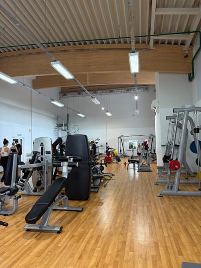 The image shows the interior of a well-lit, modern gym. The gym features a wooden floor and a high ceiling with exposed wooden beams and modern fluorescent lighting. Various exercise machines, including weight machines and benches, are arranged throughout the space. Several people are visible, engaged in activities such as working out or walking. In the background, a spiral staircase leads to an upper level. The environment appears clean and well-maintained, indicative of a functional fitness facility. The gym is located in Podkletnoe, Russia.