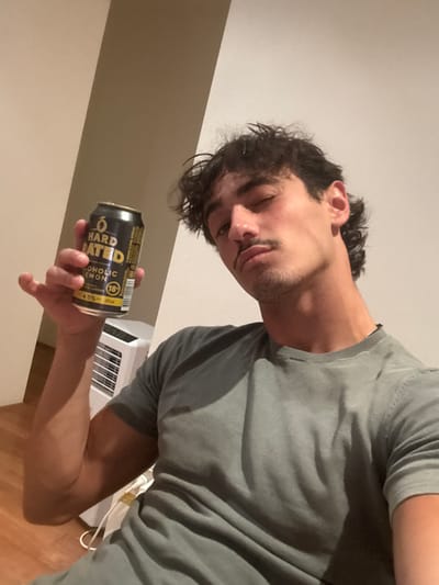 A young adult male, with dark, wavy hair styled in a mullet and a mustache with light stubble, poses for a photograph indoors in Bondi, Australia. He is wearing a greenish-grey short-sleeved t-shirt. His right hand holds a black and yellow beverage can labeled "HARD RATED ALCOHOLIC LEMON," indicating "4.5% 375ml" and "18+." His head is tilted slightly, and he looks towards the camera with a neutral expression. The background consists of plain light-colored walls with a visible architectural corner and a white vertical appliance with horizontal vents. A light-colored wooden floor with a white electrical cord is visible in the lower left.