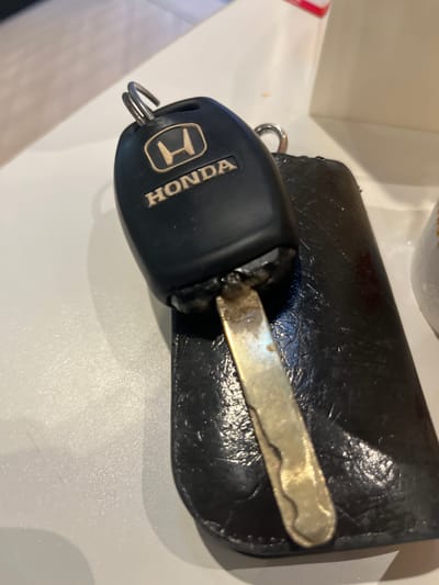 A close-up view of a black Honda car key lying on a light-colored surface next to a black leather key case. The key is attached to a key ring. The word "HONDA" and the Honda logo are visible on the key fob. The photograph was not taken in Bencongan Indah, Indonesia.