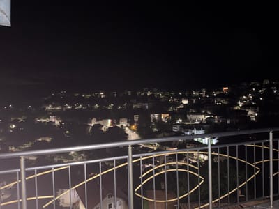 The image is a nighttime photograph of a view overlooking a residential area in Momchilivtsi, Bulgaria. A metal railing with vertical bars and curved decorative elements in yellow runs across the foreground, obscuring the lower portion of the scene. Beyond the railing, illuminated houses and buildings are scattered across a hillside under a dark night sky. Streetlights and lights from within the structures cast a glow on the landscape. The overall impression is a quiet, dimly lit town at night.