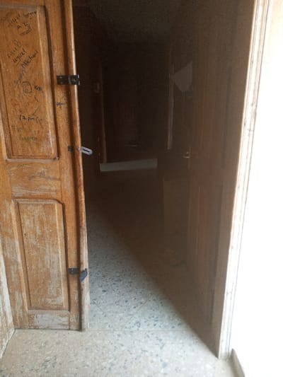 The image depicts an interior scene within a building in Oko Erin, Nigeria, looking through a partially open doorway into a dimly lit hallway or room. On the left, a light-brown wooden door, appearing aged and worn, is open approximately 45 degrees. This door features panel construction and is covered with multiple dark handwritten markings, including names such as "Fufel S. L", "Michael", "Kelly", and "Grace", along with numbers and other symbols like "12 pm". It is equipped with two black metal latches and one silver-colored hook-and-eye latch. The floor in the foreground and extending into the visible interior is covered with light-colored, speckled square tiles. A sharp, diagonal shadow line crosses the tiled floor from the bottom left to the upper right, separating a brightly illuminated area on the right from a less lit area closer to the camera. To the right of the main view, a brightly lit doorframe or wall is visible, indicating a significant light source. The area beyond the open wooden door is largely obscured by darkness, with minimal detail discernible. A faint, light-colored object, possibly a piece of paper or fabric, is visible on a wall deep within the shadowed interior, to the right. No individuals, actions, or direct interactions are present in the scene.