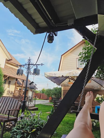 In Koledino, Russia, a low-angle perspective captures a tranquil backyard scene. The foreground is dominated by the dark wooden structure of a swing, with a person's bare legs and feet resting on it, suggesting relaxation. The midground reveals a lush green lawn interspersed with plants and flowers. A wooden bench sits to the left, positioned in front of a house with a light-colored siding and a brown roof. An ornate black lamppost with two decorative hanging lamps and a bird feeder stands between the bench and the house. In the background, two large umbrellas, branded "Rich", are set up, and a brick wall forms part of the property's boundary. The sky above is a clear blue with scattered white clouds. The overall impression is one of a peaceful summer day in a residential area.
