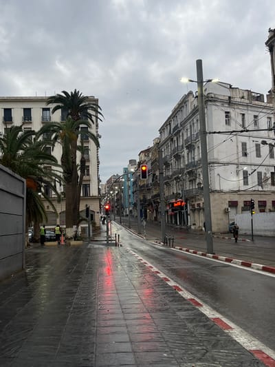 The image captures a wet, urban street scene in Oran, Algeria, under an overcast sky, indicating recent rain. The view looks down a slightly sloping street, sparsely populated.
On the immediate left, a portion of a modern, dark grey wall is visible. Behind it, a multi-story building with a white facade, dark window shutters, and balconies stands. Several tall palm trees are situated on the sidewalk in front of this building. Two individuals, wearing high-visibility yellow vests, are positioned on the left sidewalk, near a dark parked car.
The street itself, wet and reflecting light, shows a prominent red traffic light signal in the center-left. Another red traffic light is visible further down the street on the right. The road surface appears to be dark asphalt, delineated on the right by a red and white striped curb, likely indicating a dedicated tram or bus lane. Overhead, numerous black wires crisscross above the street, suspended from poles equipped with modern LED streetlights.
Along the right side of the street, a continuous row of multi-story buildings extends into the distance. These structures are predominantly white or light-colored, featuring multiple balconies and visible weathering. Ground floors house various commercial establishments, with one sign clearly displaying "RESTAURANT" in red letters. Further down, some buildings exhibit blue-toned facades.
In the midground on the right, a lone individual, dressed in a dark, long coat, walks away from the camera on the street's right side, near the red and white curb. The dark grey tiled sidewalk in the foreground is wet and reflects the red glow of the traffic lights. The overall environment suggests a quiet, possibly early morning or late afternoon, amidst damp conditions.