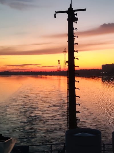 The image depicts a sunset over a body of water, likely the Volga River, near Konakovo, Russia. In the foreground, a silhouetted utility pole with climbing rungs is prominent. The sky displays a gradient of colors from pale blue at the top to vibrant orange and yellow near the horizon. In the distance, the silhouette of a city or town is visible along the riverbank, with illuminated buildings and power lines. The water surface is rippled and reflects the colors of the sunset. The overall scene conveys a sense of twilight and industrial landscape.