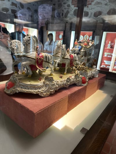 The image shows a close-up, eye-level shot of a highly ornate porcelain figurine of a horse-drawn carriage on display. The figurine features four white horses, each adorned with red and gold harnesses, pulling a detailed carriage carrying several figures dressed in historical attire. The carriage itself is elaborate, with gold trim and decorative elements. The figurine rests on a decorative, white and gold base, with red heart-shaped accents and miniature roses. This display is situated on a textured, reddish-pink velvet-covered platform, which is itself placed on a glossy, light-colored surface. In the background, reflected in a glass enclosure, are blurred figures of people and display cases containing other porcelain items, suggesting the setting is a museum or exhibition. The architectural elements of the enclosure, including arched stone walls, are also visible in the reflection. This figurine is on display at the Trakai Island Castle Museum in Trakai, Lithuania, which houses a collection of historical artifacts and decorative arts.