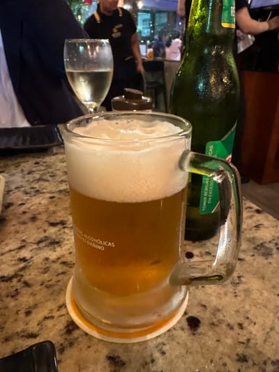 The image shows a close-up of a beer mug filled with amber-colored beer and topped with a thick layer of foam. The mug has a handle and is placed on a granite-like countertop. To the right of the mug is a condensation-covered green glass bottle of beer, with a partially visible label. In the background, out of focus, are several people and an outdoor setting, possibly a restaurant or bar, with tables and chairs. The ambient lighting suggests it is daytime or early evening. The text on the beer mug, in white, reads "IS ALCOHÓLICAS" and "ES DAÑINO," which translates to "ARE ALCOHOLIC" and "IS HARMFUL." The label on the beer bottle contains the words "TOMAR BEBIDAS" and "EN EXCESO," translating to "DRINK BEVERAGES" and "IN EXCESS." Based on the presence of these specific warning messages and the typical cultural context of such establishments, this scene is highly likely to be in Magdalena del Mar, Peru, a district known for its social venues and restaurants.