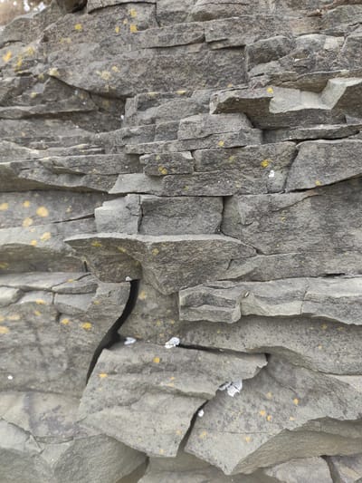 The image shows a close-up, detailed view of a layered rock formation. The rock is primarily gray with visible strata and fissures, suggesting a natural geological feature. Small flecks of yellow and white are scattered across the rock surfaces, possibly lichen or mineral deposits. The texture is rough and fractured, with sharp edges on many of the rock pieces. The lighting is even, indicating an overcast day or shaded environment. There are no people or discernible objects other than the rock itself. The location is identified as Russky Island, Russia, implying this is a natural rock face or structure found on the island, possibly near the coast or a cliffside.