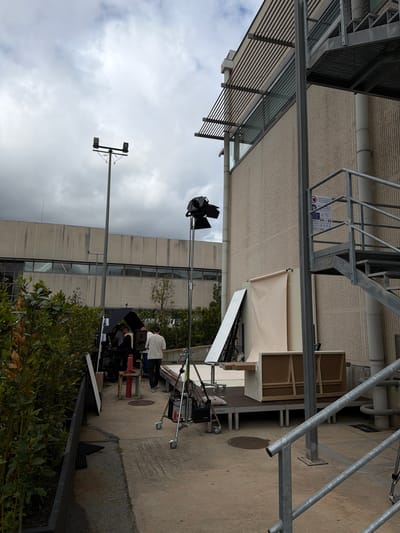 The image shows an outdoor film set. A large industrial building with multiple rows of windows is visible in the background, with film equipment set up in front. This includes lights, reflectors, and cameras on tripods. Several people are present, appearing to be crew members working on the set. A cloudy sky can be seen. The location in the city of Fibracolor, Spain, is not evident from visual cues.