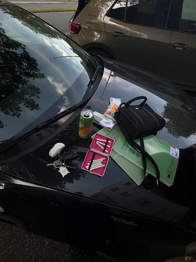 This image displays a close-up, overhead view of items placed on the hood of a black car. The scene appears to be outdoors, with reflections of trees and clouds visible on the car's surface, suggesting it is daytime. The items include a small black bag, a green folder or notebook with German text, a can of what appears to be a beverage, a pack of cigarettes, and two red cigarette cases, one filled with cigarettes. A set of keys with several keychains, including one shaped like a map of Kosovo and a miniature white sneaker, are also present.
To the left of the car, another vehicle, light-colored and with the word "GOAL" visible on its side, is partially in frame. The surrounding environment includes a road and grassy area, indicating an urban or suburban setting. The overall impression is that someone has placed their belongings on the car, possibly while stopped or taking a break. The German text on the notebook includes phrases like "Die neue Linie 1" and "EXTRAKT," and the cigarette pack also has German text. The presence of a beverage can and cigarettes suggests a casual, possibly brief, pause in an activity. There are no people visible in the frame.