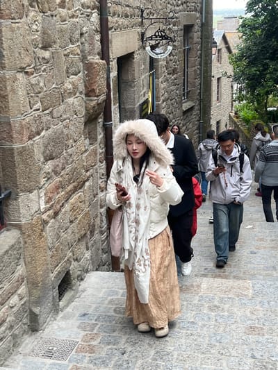 The image shows a group of people walking up a stone staircase in a medieval town. The architecture suggests a historic location, likely Le Mont-Saint-Michel, France, given the distinctive stone buildings and the steep, winding pathway. The woman in the foreground, wearing a white puffer jacket with a fur-lined hood and a long, flowy skirt, is looking down at her phone as she walks. She is holding a red object, possibly another phone, in her right hand. Behind her, other individuals are also ascending the stairs. A man in a light grey jacket and blue jeans is looking at his phone. The stone walls on either side are constructed from large, rough-hewn stones, characteristic of ancient structures. A sign hanging from the wall on the left reads "LIBRAIRIE DU SANCTUAIRE", indicating a bookstore. The lighting suggests an overcast day.