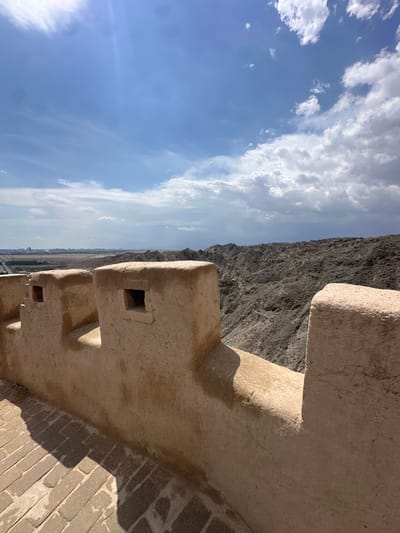 This image shows a section of an ancient defensive wall, likely part of the Jiayuguan Pass, also known as the "First Great Fortification Under Heaven," located in Jiayuguan City, Gansu Province, China. The wall, constructed from rammed earth and possibly adobe, features crenellations and small rectangular openings, suggestive of watch posts or arrow slits. The weathered texture of the wall indicates its age and exposure to the elements. In the foreground, a cobblestone pathway or walkway is visible, with distinct shadows cast by the wall structures, suggesting a bright, sunny day. Beyond the wall, the landscape transitions to arid, rocky terrain, characteristic of the Hexi Corridor region, with rolling hills and rugged mountains stretching towards the horizon. In the far distance, a hazy, urban or semi-urban area with industrial structures can be observed, with a road leading towards it. The sky above is a dynamic mix of blue and white clouds, with sunlight filtering through, creating a sense of depth and atmospheric perspective. There are no people visible in the frame.
