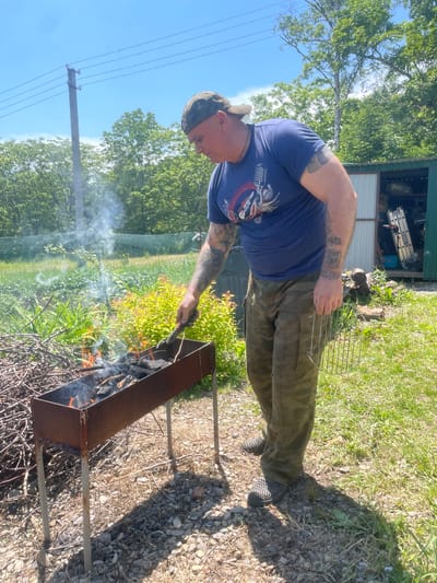 A man wearing a blue t-shirt, camouflage pants, and a camouflage baseball cap is standing outdoors in what appears to be a yard. He is holding a metal poker, tending to a fire in a rusted metal barbecue grill filled with charcoal and burning wood. Smoke is rising from the grill. To the left of the grill, a pile of dry branches is visible. In the background, there is a green fence, lush green foliage, trees, a utility pole with wires, and a small green shed with a ladder inside. The ground is covered with a mixture of grass, gravel, and dry debris. The sky is blue and clear. This scene is located in Yasnoe, Russia.