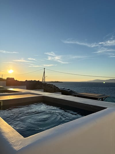 The image displays a coastal sunset scene on the island of Mykonos, Greece, offering a panoramic view of the Aegean Sea. In the immediate foreground, two elevated, rectangular pools or hot tubs are visible. The closest pool, on the right, contains dark blue water with surface ripples, suggesting active jets or recent disturbance. It features white coping and dark tiled interior edges. Adjacent to it, a second, partially visible pool is situated to the left. The surrounding structures are rendered in white, consistent with Cycladic architecture.
Beyond the pools, the midground reveals a rugged, rocky coastline descending towards the sea. A dark utility pole with overhead wires is centrally positioned against the sky. Two white vehicles are parked on a lower terraced area near the water's edge. A wooden platform or deck structure extends towards the sea on the right.
The background is dominated by the expansive, calm Aegean Sea, with distant landmasses visible on the horizon to the right. The sun is a bright, golden orb positioned on the left horizon, indicating sunset. The sky transitions from warm yellow and orange near the sun to a clear light blue overhead, with scattered wispy white clouds. No individuals are present in the scene.