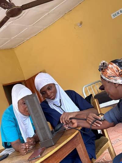 The scene depicts three individuals indoors. Two individuals, both wearing white headscarves, are positioned at what appears to be a wooden desk or examination table. One wears a blue top, and the other wears a navy blue top with a white undershirt. The person in blue is holding their hands together, seemingly awaiting or receiving medical attention. The second individual with a navy blue top has a stethoscope around their neck and is holding medical equipment, possibly a sphygmomanometer to measure blood pressure. The third individual, wearing a patterned headscarf, is above and slightly behind, positioned as if taking a blood pressure reading on the arm of the individual sitting in navy blue. The setting could be a clinic or medical examination room. A sign that says "Sick B" is visible on the wall. The lighting is natural. Based on the information available, it is impossible to verify its location in Madangala, Nigeria.