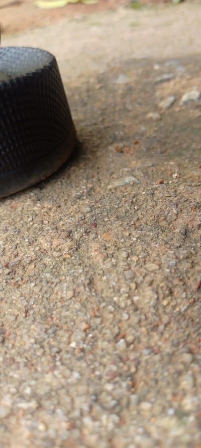 The image shows a close-up, low-angle view of a textured, black, thimble-like object resting on a granular, earthy surface. The surface is composed of small pebbles, sand, and dirt, with variations in color from light beige to reddish-brown. The object is partially in focus, revealing a diamond-shaped pattern on its exterior. The background is out of focus, showing hints of blurred greenery and a lighter, possibly dirt or gravel, area. The scene appears to be outdoors, on the ground. There is no information available to specifically place this scene within the city of Baissa, Nigeria.