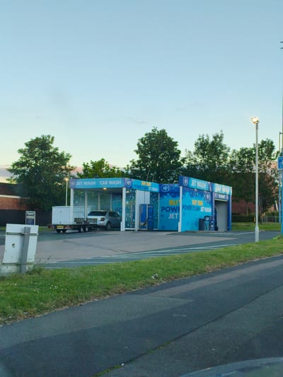In Sandwell, United Kingdom, at dusk, a car wash facility is visible. The facility is a modern, open-air structure with blue and white branding. The words "JET WASH" and "CAR WASH" are prominently displayed on the building. A silver sedan is positioned within the wash bay, and a trailer is attached to the rear of a white box truck parked nearby. The area surrounding the car wash is paved, with a grassy verge and a road in the foreground. A tall lamppost with a lit bulb stands to the right of the car wash, illuminating the scene. Trees form a backdrop to the car wash, and a brick building is visible in the distance. The sky is a pale blue, indicative of twilight.