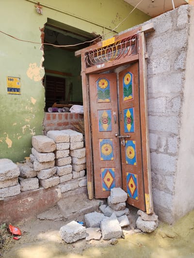 An exterior scene in Pedda Kammavaripalli, India, displays a partially demolished or under-construction building. The left section features a light green wall with significant peeling paint and a large, irregular vertical opening revealing a darker green interior wall, a brown window frame, and a ceiling fan. A yellow and blue poster, marked "198" and "BSNL," is affixed to the green wall. A thin brown cable traverses the upper portion of this wall. Inside the opening, on a raised brick platform, are plastic bottles, a white bag, and a broom.
To the right, a detached brown wooden double door, decorated with multiple colorful stylized floral patterns in yellow, blue, red, and green, leans against an unfinished light grey concrete block wall. The door is secured by a silver bolt latch and has a silver handle on its left leaf. Above the door, a wooden lintel supports a red and yellow decorative metal grille, with labels "W-222" and "W-389" attached. The bottom right edge of the door is propped up by several rough grey concrete blocks.
On the ground, a large stack of light grey, coarse concrete blocks is piled against the base of the green wall. Additional scattered blocks lie on the dirt and sand ground, which also contains a red plastic wrapper and dry leaves. Yellowish sand or fine gravel is visible near the base of the concrete block wall. No individuals are present in the scene.