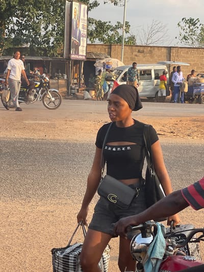 The image depicts a bustling street scene, likely in an urban or semi-urban area, during daylight hours with a clear sky. The photo is oriented ninety degrees clockwise, causing elements to appear on their sides.
In the foreground, a young woman with dark skin stands on a paved road, looking towards the left side of the frame with a slight smile. She is wearing a black head covering, a short-sleeved black crop top with a cutout revealing part of her midriff, and dark shorts. The crop top has white stylized text on the chest, which appears to spell "DESSERT". A black shoulder bag with a gold C-shaped clasp is slung across her body. Her right hand holds the handle of a checkered (black and white plaid) rolling bag or suitcase, which rests on the ground beside her.
To her immediate right (viewer's left when rotated correctly), the partial figure of another person is visible, primarily their arm and hand. This person, also with dark skin, is sitting on a red and silver motorcycle, holding the handlebars. They are wearing a red and black striped shirt. The woman appears to be either riding with or waiting for this person.
The midground and background reveal more of the street activity. On the left side of the image, there is a large billboard displaying images of men in suits, likely a political or commercial advertisement. Part of the text "PROFESSOR" is visible on this billboard. Below the billboard, another motorcycle is parked, and a person is seen standing near it. Behind these elements, there are structures that appear to be small shops or stalls with corrugated metal roofs and brick walls, surrounded by green foliage and trees. A utility pole stands upright.
In the center-right background, a light-colored, older model mini-bus or van is present, surrounded by several people. Some individuals are standing near the van, while others appear to be entering or exiting it. One person inside the van is wearing a shirt with red and yellow colors, possibly resembling a flag pattern. Further in the background, more pedestrians are visible, moving along the roadside.
The road surface is a mix of asphalt and lighter, dusty patches. The overall impression is one of a lively street environment, possibly a market area or a public transport hub, under a bright sun.