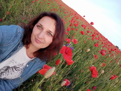 A woman with dark, shoulder-length hair and blue eyes is lying in a field of red poppies. She is wearing a denim jacket over a white t-shirt with a graphic of a cat. The field extends into the distance, with the poppies creating a vibrant red carpet against the green stems and stalks. The sky above is a pale blue with a slight gradient. The photograph appears to be taken in the village of Velyka Omeliana, Ukraine.