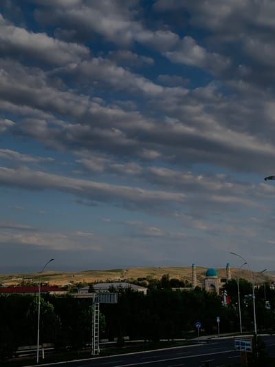 The image depicts a wide shot of Zaamin, Uzbekistan, with a focus on the sky and the landscape. A dramatic sky filled with layered clouds dominates the upper portion of the frame. Below the sky, rolling, light-brown hills form the background. In the middle ground, a mosque with a prominent turquoise dome and two minarets stands out, surrounded by lush green trees. Some residential buildings with red and light-colored roofs are visible to the left of the mosque. A road runs through the foreground, with streetlights and traffic signs visible. The overall scene suggests a partly cloudy day in a semi-arid urban or semi-urban environment.