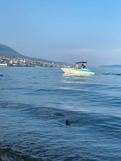 In Milvignes, Switzerland, a motorboat with two occupants cruises across a body of water under a clear sky. The boat, white with a turquoise stripe and a dark canopy, is moving from left to right, leaving a visible wake. In the background, a densely populated city sprawls along the base of a green, tree-covered mountain. The water exhibits gentle ripples, and along the left edge of the frame, the rocky shoreline is visible.