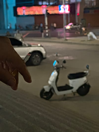 A close-up shot focuses on a person's hand holding a lit cigarette. In the background, a white electric scooter is parked on a street. Further in the distance, a white car is visible, along with blurred neon signs displaying red and blue lights. The scene appears to be an urban environment, potentially in Perímetro Urbano Barranquilla, Colombia, at night, given the artificial lighting and parked vehicles.