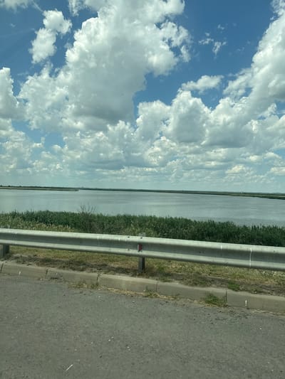 This image captures a roadside view, likely from a moving vehicle, overlooking a body of water and a vast sky. The scene is dominated by a wide, calm expanse of water, possibly a river or lake, which stretches towards a distant, hazy horizon. Along the near edge of the water, dense green vegetation, likely reeds or tall grass, forms a natural barrier.

To the right of the vegetation, a grey asphalt road runs parallel to the water, bordered by a concrete curb. A metal guardrail, made of two horizontal silver bars supported by vertical posts, separates the road from the grassy embankment. The guardrail shows signs of wear, with some scuffs and possibly reflections of red reflectors. The road surface itself is speckled with small stones and appears to be a typical paved highway.

Above, the sky is a vibrant blue, filled with large, puffy white cumulus clouds. These clouds are dynamic, with some appearing bright and sunlit, while others are softer and more diffused, suggesting a bright, partly cloudy day. The overall impression is one of a scenic drive, possibly in a rural or semi-rural area.

The time of day appears to be daytime, given the brightness of the sky and the lighting on the clouds. The weather seems pleasant, with clear skies and sunshine indicated by the illuminated clouds. There are no people visible in the image, and the focus is on the natural landscape and the roadside infrastructure. The location is identified as Krasnyi, Russia. No visible text is present in the image.