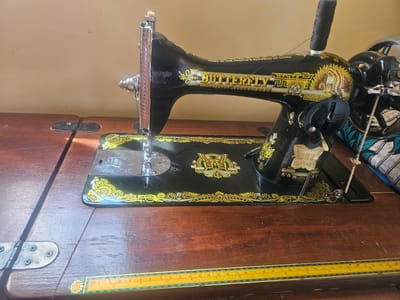 This is a close-up, eye-level shot of a black, antique sewing machine with gold decorative elements, primarily the "Butterfly" brand name. The sewing machine is positioned on a wooden table, which appears to be part of a sewing machine base or cabinet. The wood is dark and shows some grain and wear. A ruler with cm and inch markings is visible along the bottom edge of the table. In the background, the wall is a plain, light beige color. The scene is lit by natural light, casting a shadow from the sewing machine onto the table. The sewing machine itself is detailed, with a spool of black thread on top, a needle and presser foot, and various levers and dials. A piece of patterned fabric, with teal and white designs, is visible on the right side of the image, behind the sewing machine. The specific location in the city of Masaka, Nigeria, cannot be determined from the visual information provided in the image.