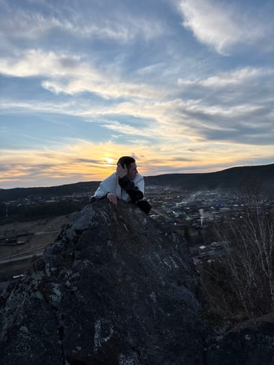 A person is sitting on a large, dark-colored rock at the edge of a cliff. The person is wearing a white hooded sweatshirt and dark pants, with their arms crossed and head resting on their hands. The setting sun is directly behind the person, casting a warm glow on the sky and creating a silhouette effect. In the background, there is a village or town, possibly Невон, Russia, nestled in a valley with a mountain range in the distance. The sky is filled with white and grey clouds.