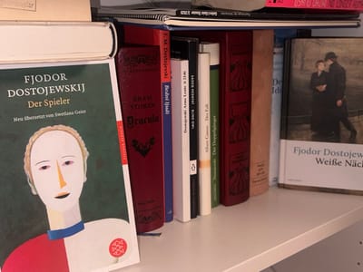 The image shows a bookshelf filled with various books. Dominating the left side is "Der Spieler" by Fjodor Dostojewskij, with a striking cover illustration. Adjacent to it is Bram Stoker's "Dracula," its cover featuring a bat. Other books include "Bedni ljudi" by F.M. Dostojevski, Sadeq Hedayat's "Blind Owl", Albert Camus' "The Fall", and a book entitled "Der Doppelganger" by Dostoevsky. On the right, "Weiße Nächte" by Fjodor Dostojewskij stands out, along with an assortment of other titles, including "De Idi" that are partially obscured. Some magazines or journals can be seen on the top shelf.