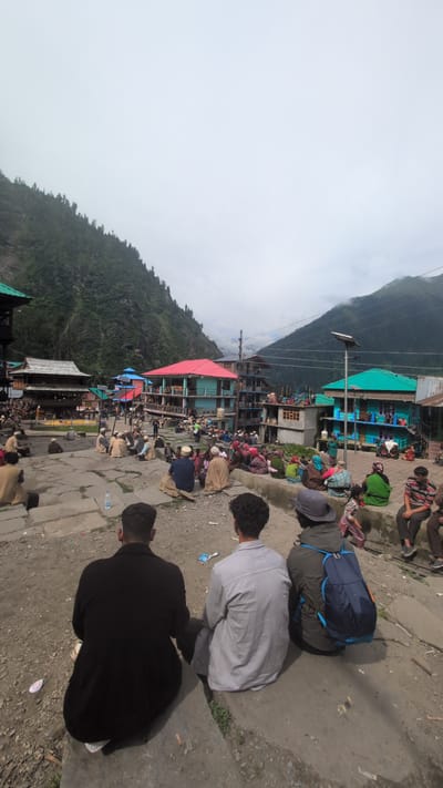 The image depicts a view of the village of Malana, India, with a focus on a gathering of people in an open area. In the foreground, three individuals are seated with their backs to the camera, observing the scene. The middle person is wearing a light-colored shirt and a dark hat, with a blue backpack on their back. To their left, another person is in a black jacket, and to their right, a person is in a light gray shirt.
The middle ground features a densely populated area with many people seated on the ground and on raised platforms. They are dressed in traditional clothing, with many women wearing colorful headscarves and shawls. The buildings in this area are colorful, with prominent red and blue roofs, indicating a village setting. The architecture suggests a traditional style, with wooden elements and multiple stories.
In the background, steep, tree-covered mountains rise up, partially obscured by clouds, suggesting a mountainous region. The sky is overcast, with a grey, cloudy atmosphere. The ground in the gathering area appears to be paved with stone or concrete, with some dirt and debris present. Power lines are visible, strung between poles, indicating the presence of electricity infrastructure. The overall scene suggests a community event or gathering in a remote mountain village.