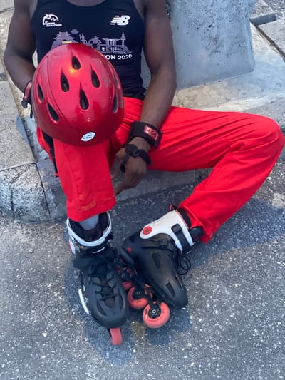 The image shows a person sitting outdoors on a concrete surface, possibly a step or low wall. The person is wearing red pants, a black tank top with a New Balance logo and a "SEOUL MARATHON 2020" graphic, black inline skates with white and red details, a red helmet, and wrist guards. The location is not explicitly identified as Sangotedo, Nigeria, but given the clothing and skin tone of the subject, it may be accurate.