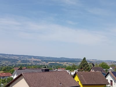 The image shows a view of the city of Ottenbach, Switzerland, on a clear day. The foreground is dominated by the rooftops of several houses with brown, tiled roofs. One house in the lower right corner has a bright yellow facade and a brown tiled roof. Further in the background, there are more houses with various roof colors and architectural styles, nestled amongst green trees. Beyond the residential area, a rolling landscape of fields and forests stretches towards the horizon. The sky is a pale blue with wispy white clouds. The scene suggests a peaceful, residential suburban environment with a view of the surrounding countryside.