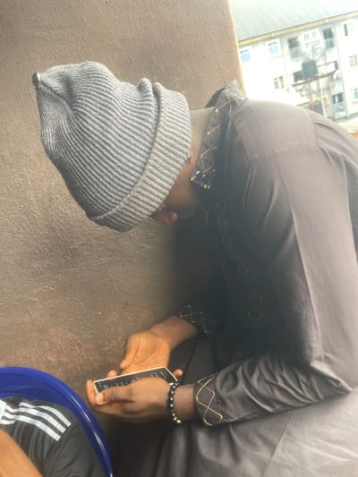 A person in a gray robe and matching beanie bows his head, eyes closed. The robe has an ornate gold-colored pattern near the neckline and wrists. The person holds a white smartphone in his left hand, and a black beaded bracelet is visible on the right wrist. Parts of an exterior building are seen outside, and a person with a striped Adidas outfit is partially visible at the bottom-right corner.