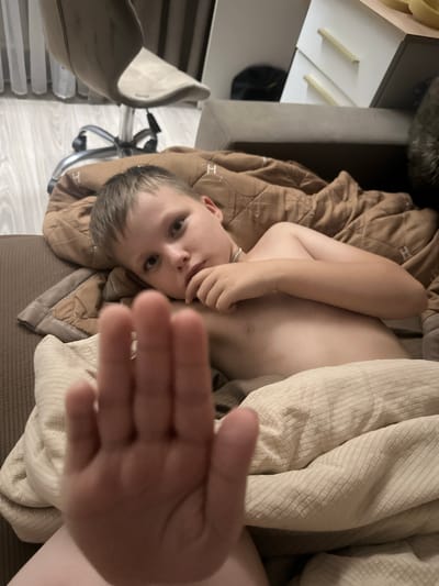 The image shows a young person lying in bed, covered by blankets. The person is looking towards the camera with one hand held up, partially obscuring their face. They appear to be indoors, with a chair and what looks like a desk or shelf visible in the background. The lighting suggests it might be daytime, though the indoor setting makes it difficult to determine the exact time of day or weather conditions. The overall mood of the image is informal and personal, possibly capturing a candid moment. There is no visible text in the image.